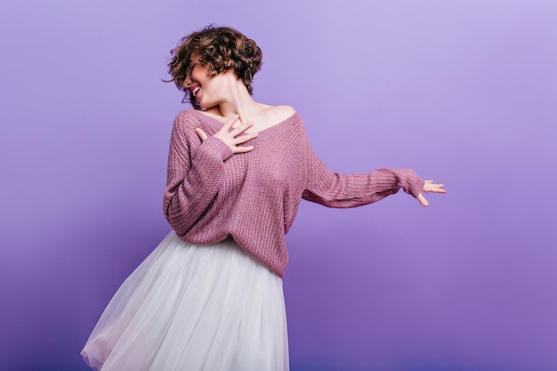 Mujer romántica inspirada con el pelo corto bailando en estudio en falda larga blanca. Chica morena alegre en suéter de gran tamaño tejido pasar tiempo en la sesión de fotos.