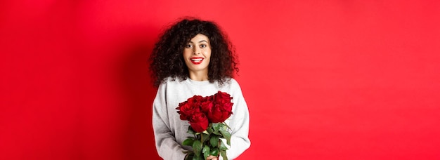 Foto gratuita mujer romántica con peinado rizado recibe ramo de rosas rojas en el día de san valentín mirando tierno