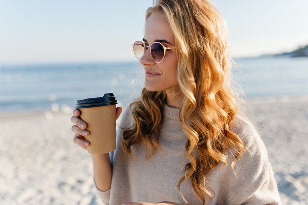 Mujer romántica con pelo ling blinde bebiendo té en el mar. Retrato al aire libre de una mujer encantadora con gafas de sol mirando al mar en la mañana de otoño.