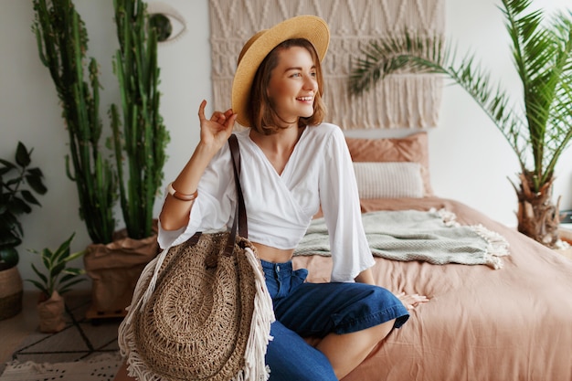 Mujer romántica con sincera sonrisa sentada en la cama, disfrutando de la mañana soleada en su elegante piso en estilo boho