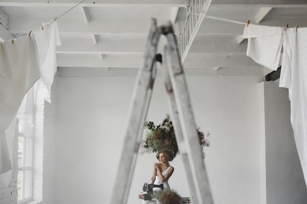 La mujer en la ropa blanca se sienta en una habitación con flores y una máquina de coser