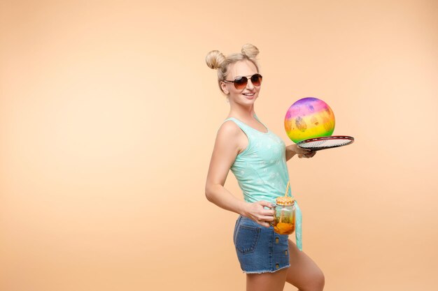 Una mujer con ropa brillante, una camiseta y pantalones cortos de mezclilla, gafas de sol, sostiene una pelota y limonada fría en su