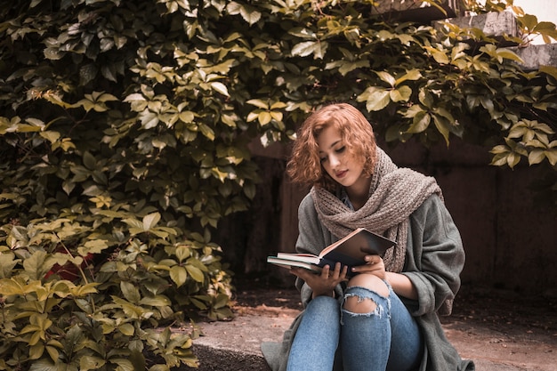 Mujer en ropa de calle sentado a bordo y leyendo atentamente el libro cerca de la planta