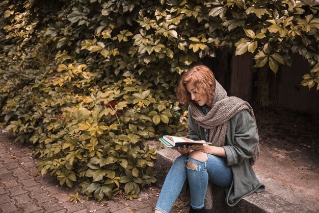 Mujer en ropa de calle sentado a bordo y leyendo el libro cerca de la planta