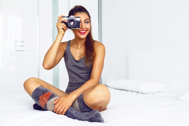 Mujer en ropa casual acogedora, sentada en una gran cama blanca y tomar fotografías con una cámara vintage