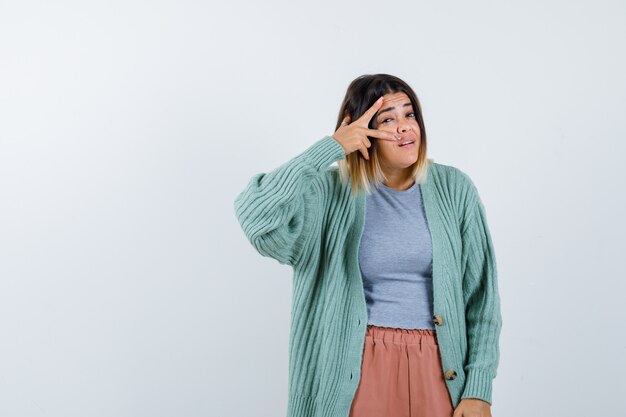 Mujer en ropa casual mostrando el signo V en el ojo y mirando confiada, vista frontal.