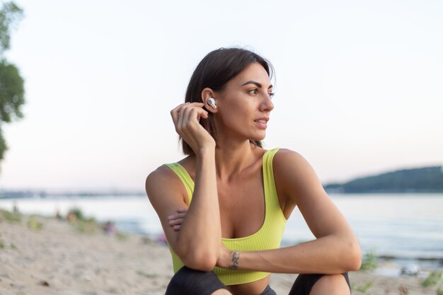 Mujer en ropa deportiva al atardecer en la playa de la ciudad descansando después del entrenamiento escuchando música en auriculares inalámbricos