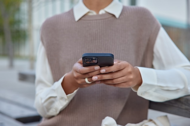 mujer con ropa elegante sostiene teléfono móvil navega por internet inalámbrico hace compras en línea se sienta en un banco afuera conectado a internet inalámbrico se comunica a distancia