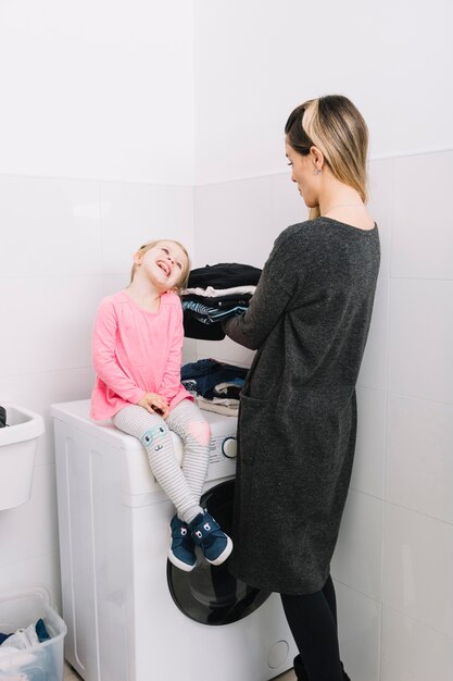 Mujer con ropa sucia mirando a su hija linda sentada en la lavadora