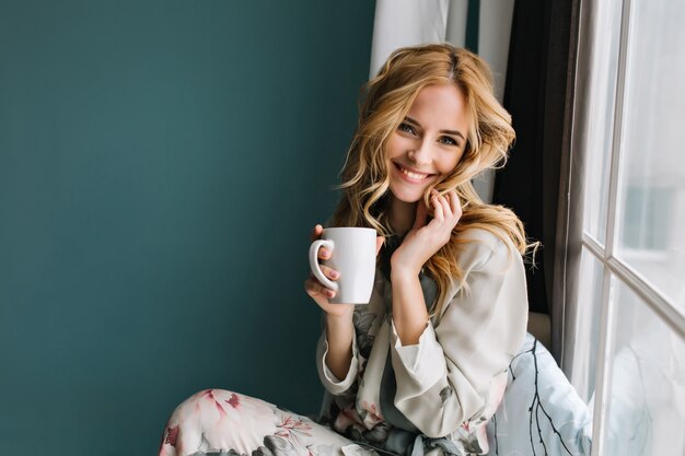 Mujer rubia alegre relajarse y sentarse en el alféizar de la ventana, sosteniendo una taza de café, té. Ella tiene cabello largo y rubio ondulado, hermosa sonrisa. Usando un bonito pijama con flores.