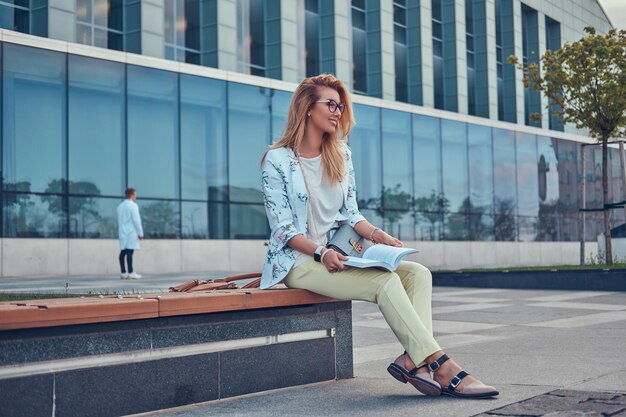 Mujer rubia alegre con ropa moderna, estudiando con un libro, sentada en un banco en el parque contra un rascacielos.