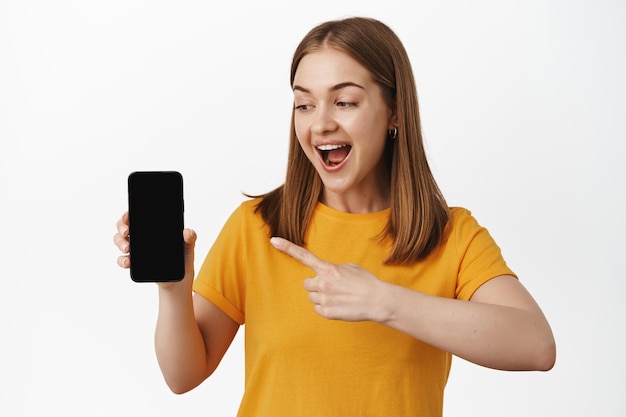 Foto gratuita mujer rubia alegre señalando con el dedo la pantalla vacía del teléfono, la interfaz de la aplicación, mostrando la venta en la pantalla móvil, recomendar la aplicación para smartphones, fondo blanco