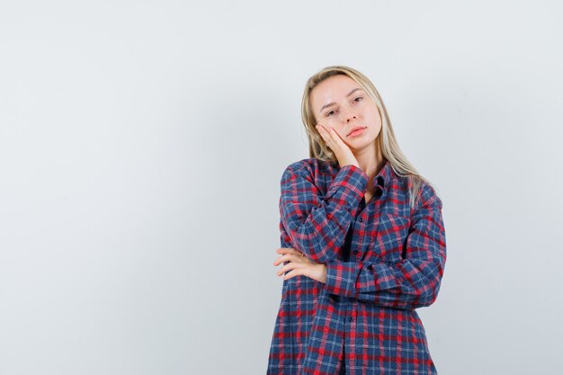 Mujer rubia apoyándose en la palma de la mano en camisa a cuadros y mirando atractiva, vista frontal.