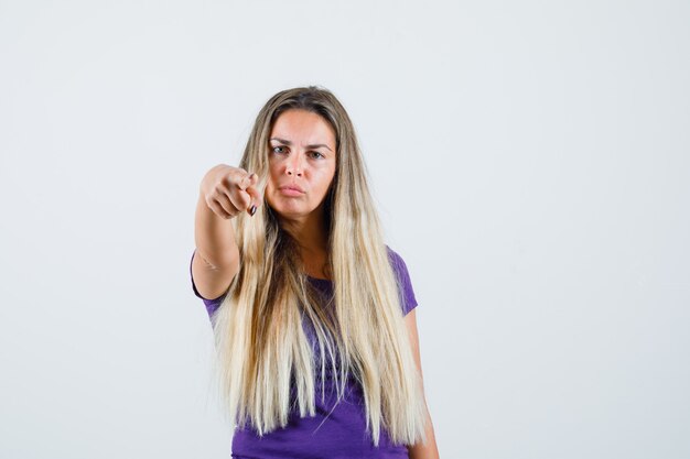 mujer rubia apuntando al frente con camiseta violeta y mirando seria. vista frontal.