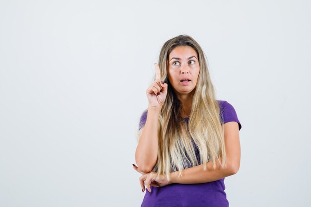 mujer rubia apuntando hacia arriba con camiseta violeta y mirando enfocado, vista frontal.