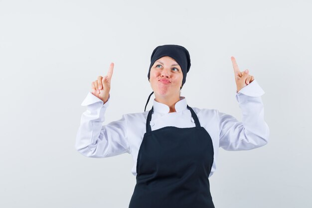 Mujer rubia apuntando hacia arriba con los dedos índices, labios curvados en uniforme de cocinero negro y luciendo bonita