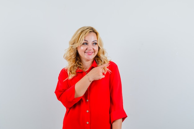 Mujer rubia apuntando a la derecha con el dedo índice en blusa roja y mirando feliz