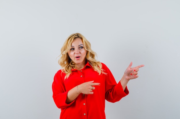 Mujer rubia apuntando a la derecha con los dedos índices en blusa roja y mirando feliz