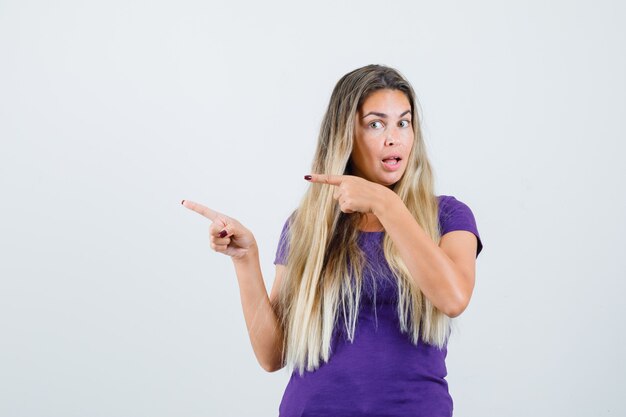 mujer rubia apuntando a un lado en camiseta violeta y mirando curiosa. vista frontal.