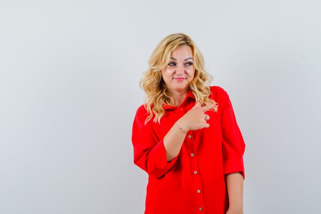 Mujer rubia apuntando hacia el lado derecho con el dedo índice en blusa roja y mirando feliz.