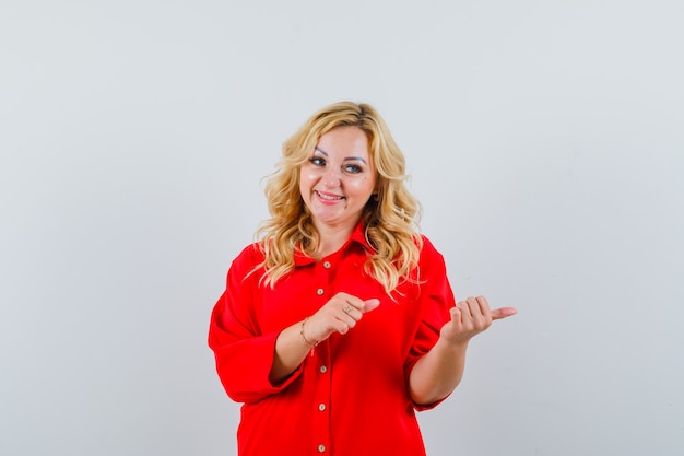 Mujer rubia apuntando hacia el lado derecho con los dedos índices en blusa roja y mirando feliz.