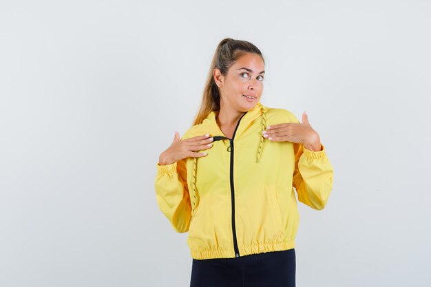 Mujer rubia apuntando a sí misma con los dedos índices en chaqueta de bombardero amarillo y pantalón negro y mirando feliz