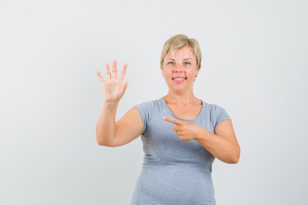 Mujer rubia apuntando a su mano y sonriendo con camiseta azul claro y mirando feliz, vista frontal.