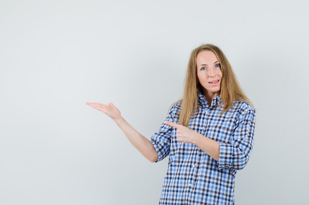 Mujer rubia apuntando a su palma extendida a un lado en camisa y mirando confiada,