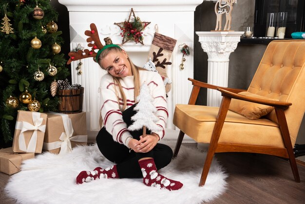 Mujer rubia con árbol de Navidad blanco posando junto a la chimenea.