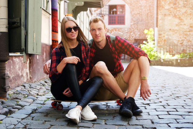 Mujer rubia atractiva con gafas de sol y su novio sentado en longboard en una calle del casco antiguo.