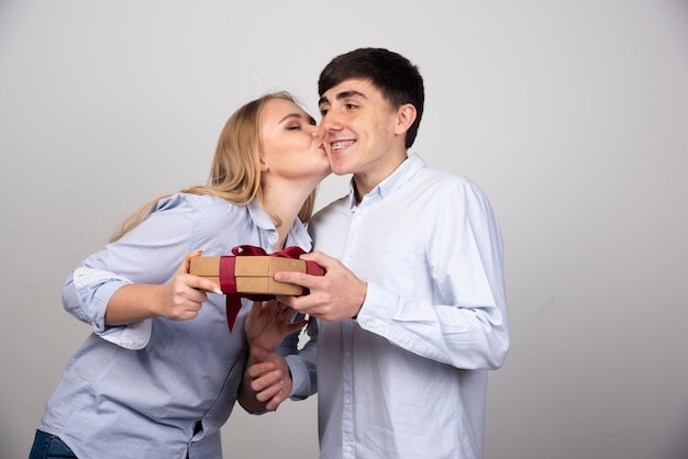 Mujer rubia besando a su novio con regalo presente contra la pared gris.