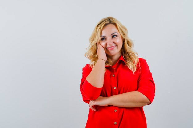 Mujer rubia en blusa roja apoyándose en la mejilla en la mano, posando a la cámara y mirando feliz,