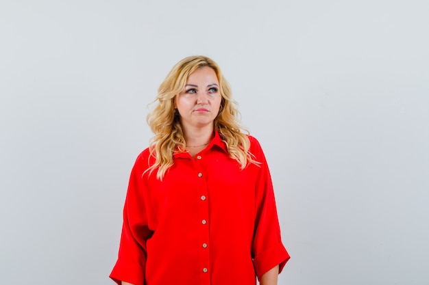 Foto gratuita mujer rubia en blusa roja de pie derecho y posando a la cámara y mirando serio