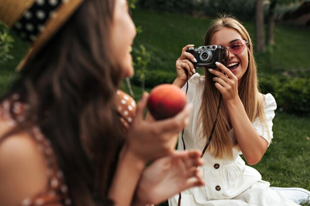 Mujer rubia bronceada con gafas de sol rojas sostiene una cámara retro y toma una foto de su novia morena rizada Las mujeres tienen un picnic afuera