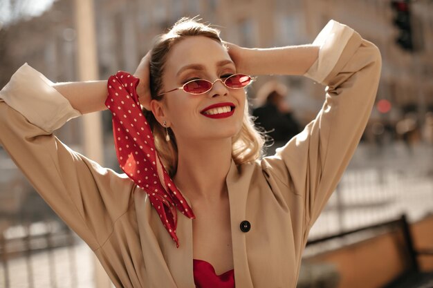 Mujer rubia de buen humor con blusa de seda de gabardina beige y gafas de sol brillantes sonríe sinceramente fuera Señora feliz con labios rojos toca el cabello