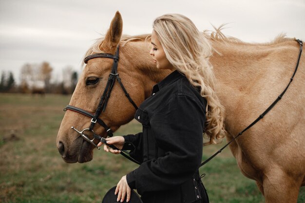 Mujer rubia y caballo marrón parados en el campo. Mujer con ropa negra. Mujer tocando el caballo.