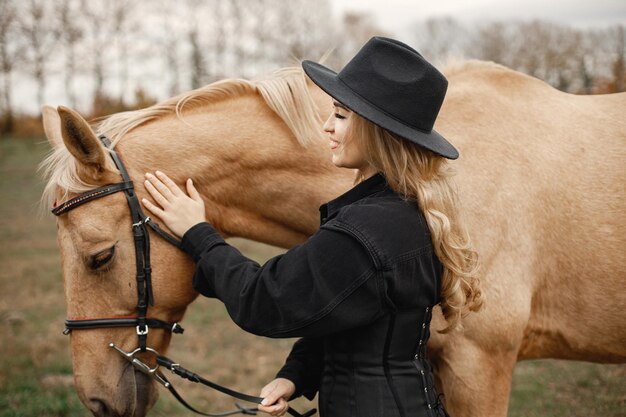 Mujer rubia y caballo marrón parados en el campo. Mujer vestida de negro y sombrero. Mujer tocando el caballo.