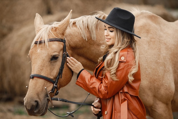 Mujer rubia y caballo marrón parados en una granja cerca de pacas de heno. Mujer con vestido negro, abrigo de cuero rojo y sombrero. Mujer tocando el caballo.