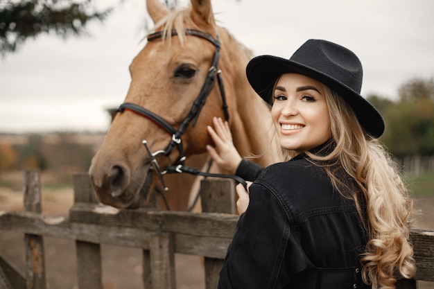 Mujer rubia y caballo marrón parados en una granja. Mujer vestida de negro y sombrero. Mujer tocando el caballo detrás de la valla.