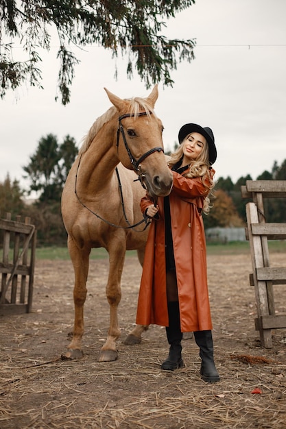 Mujer rubia y caballo marrón parados en una granja. mujer con vestido negro, abrigo de cuero rojo y sombrero. mujer tocando el caballo.