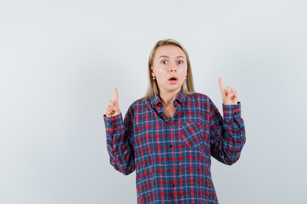 Mujer rubia en camisa a cuadros apuntando hacia arriba con los dedos índices y mirando sorprendido, vista frontal.