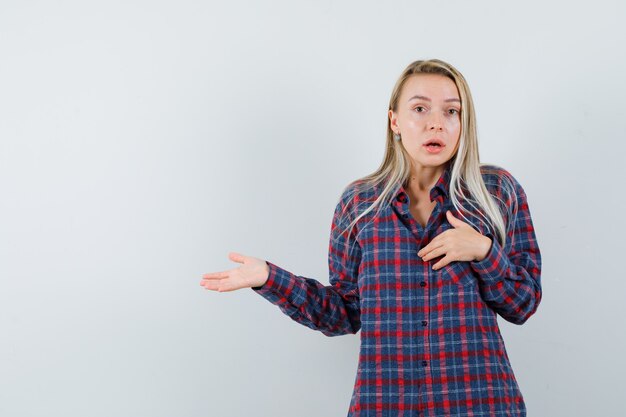 Mujer rubia en camisa a cuadros estirando las manos hacia la izquierda y mirando sorprendido, vista frontal.