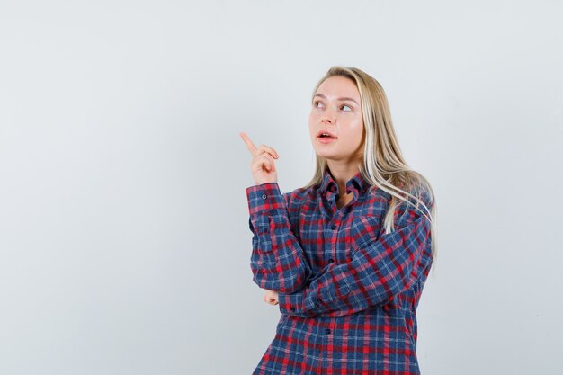 Mujer rubia en camisa a cuadros levantando el dedo índice en gesto de eureka y mirando precavido, vista frontal.