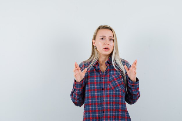 Mujer rubia en camisa a cuadros levantando las manos como para explicar algo y mirando disgustado, vista frontal.