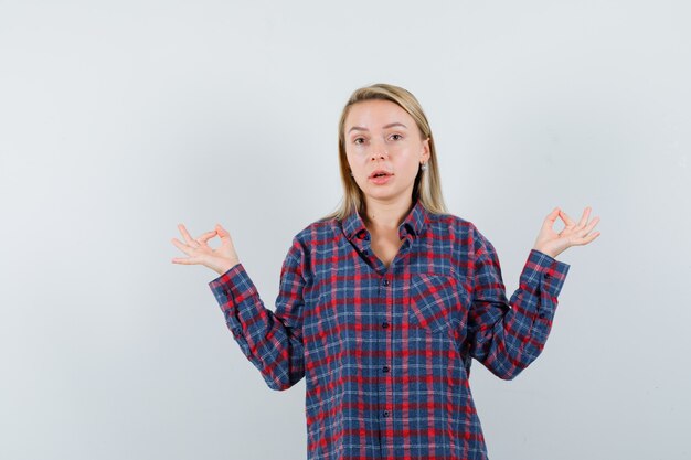 Mujer rubia en camisa de cuadros meditando y mirando sorprendido, vista frontal.