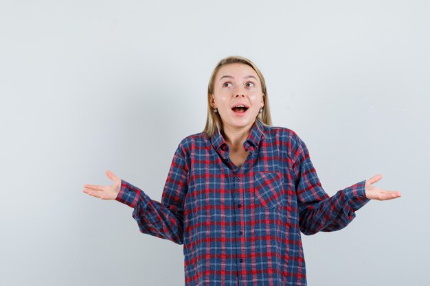 Mujer rubia en camisa a cuadros mostrando gesto de impotencia y mirando sorprendido, vista frontal.