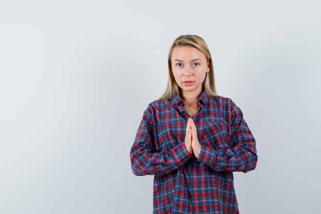 Mujer rubia en camisa a cuadros mostrando gesto de namaste y mirando enfocado, vista frontal.