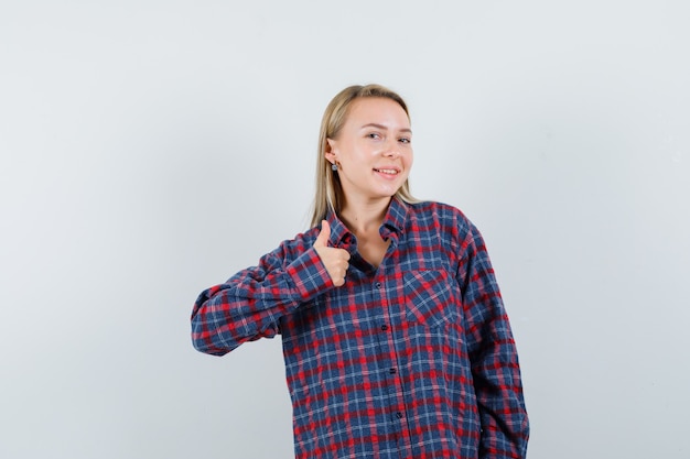 Mujer rubia en camisa a cuadros mostrando el pulgar hacia arriba y mirando optimista, vista frontal.