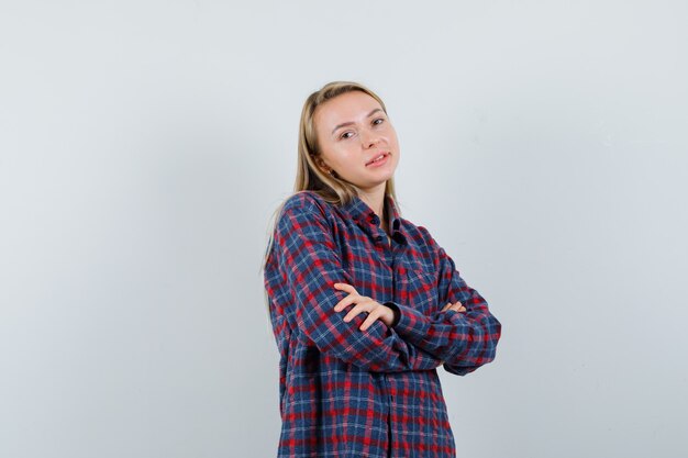 Mujer rubia en camisa a cuadros de pie con los brazos cruzados y mirando feliz, vista frontal.