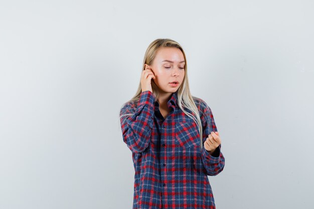 Mujer rubia en camisa a cuadros que finge hablar por teléfono mientras mira a la mano como sosteniendo algo y mirando enfocado, vista frontal.